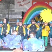 Bild vergr&ouml;&szlig;ern: Kost&uuml;mierte Oelsnitzer Kinder singen und tanzen auf der Aktionsb&uuml;hne vor einer Sonne-Wolken-Regenbogen-Kulisse.